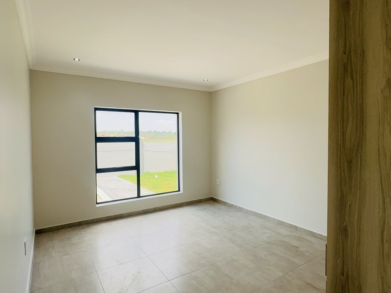 A bright, empty bedroom featuring neutral walls, tiled flooring, and a large aluminium-framed window providing natural light.