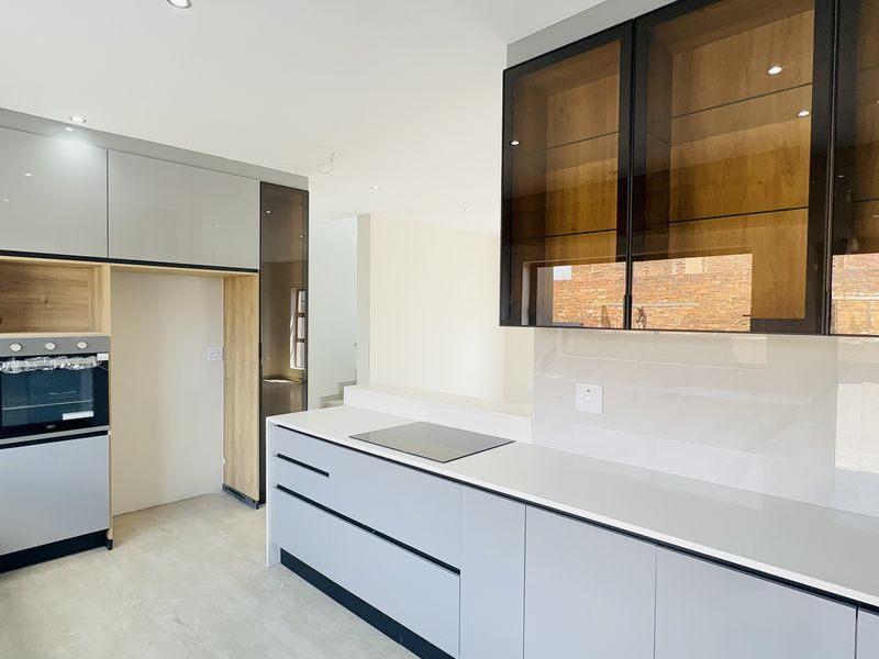 Modern kitchen featuring sleek grey gloss cabinetry, integrated oven, induction hob, quartz countertops, and stylish glass-fronted display units in a bright, open-plan layout.