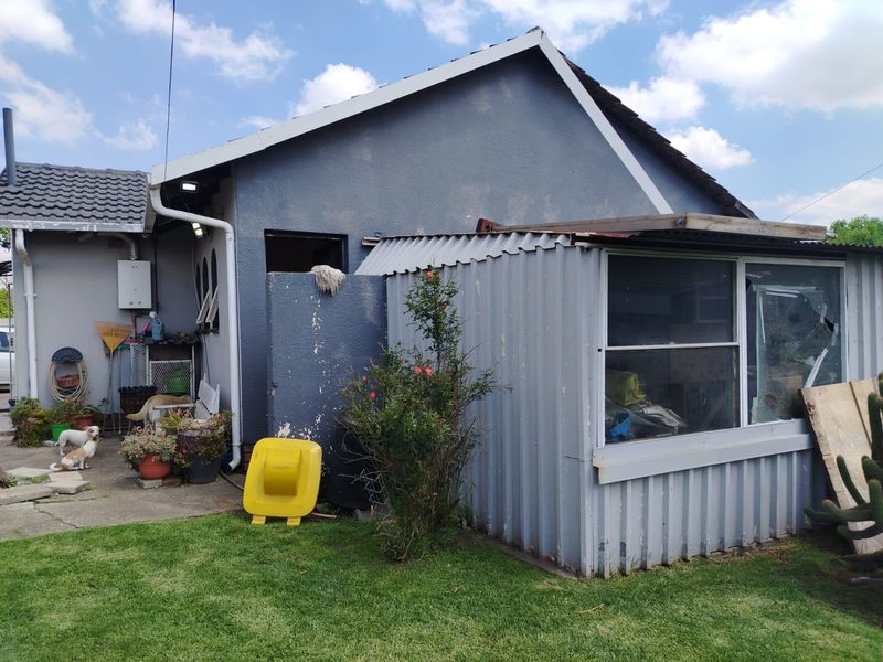 Rear view of the property showing a main structure with a corrugated metal lean-to extension, concrete patio area, and visible exterior maintenance requirements.