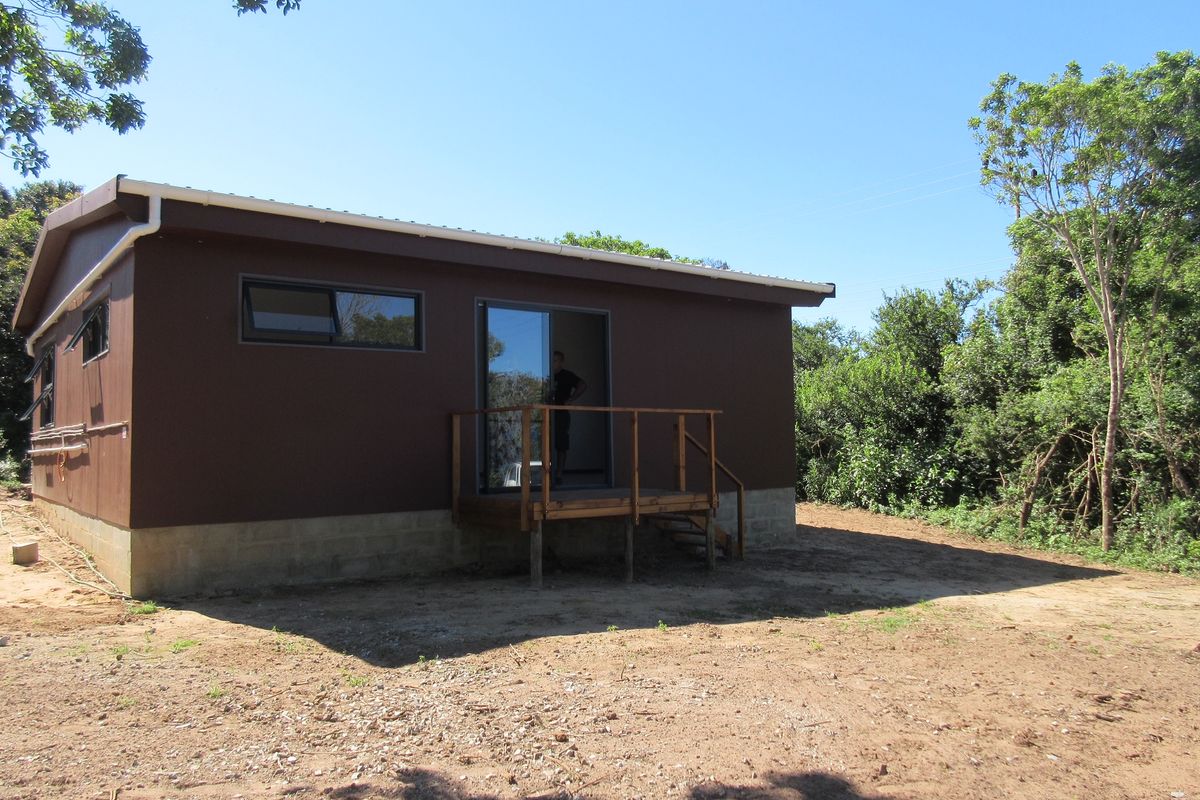 Rear view of a modern brown-painted house featuring a small timber deck, sliding glass doors, and awning windows set against a natural garden backdrop.