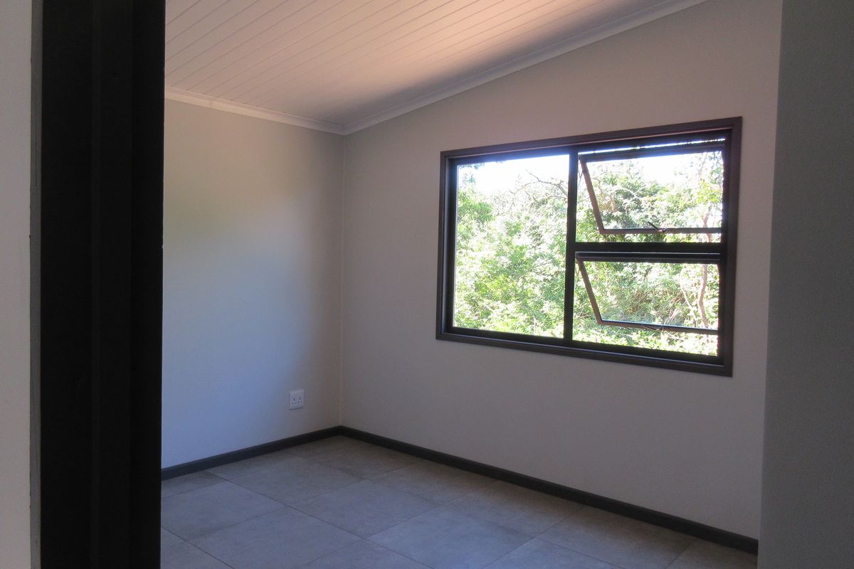 A clean, empty bedroom featuring neutral tiled flooring, a tongue and groove ceiling, and a window with views of the surrounding greenery.