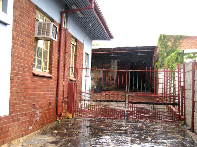Rear exterior area featuring a paved courtyard, brick facade, and a metal security gate leading to a covered structure with visible wear.