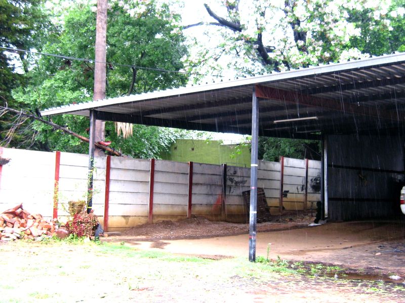 An open-sided carport structure with a corrugated metal roof and precast concrete boundary walls, showing signs of wear and poor drainage.