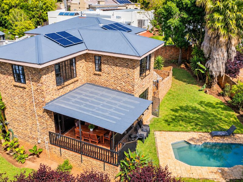 Aerial view of a double-story brick house featuring a corrugated metal roof with solar panels, a swimming pool, and a covered patio area.