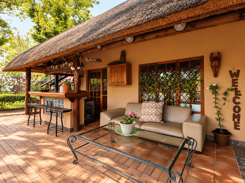 A charming covered patio featuring a thatched roof, timber support beams, and a built-in bar area, overlooking a well-maintained garden space.