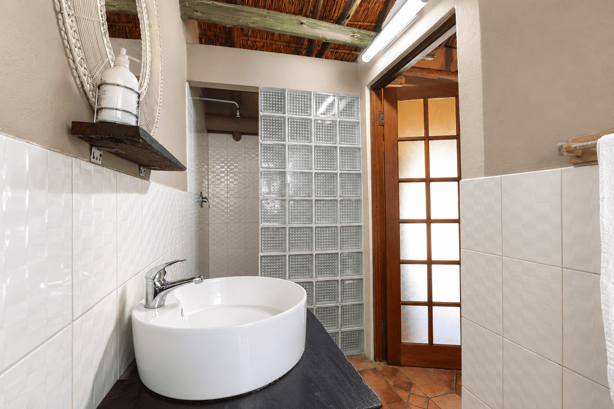 A bathroom featuring a vessel sink, glass block shower partition, terracotta flooring, and a unique thatched roof ceiling.