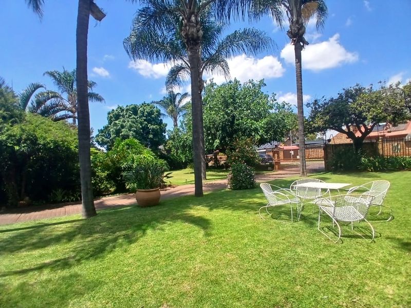 A well-maintained garden featuring a lush lawn, mature palm trees, a paved walkway, and secure perimeter walling with a metal gate.