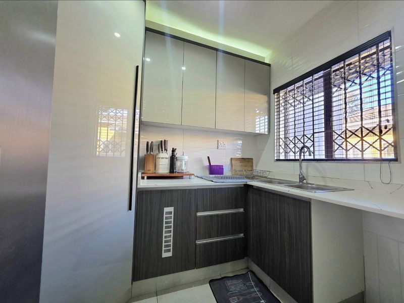 A modern, compact kitchen featuring sleek two-tone cabinetry, stone countertops, a stainless steel sink, and a window fitted with a security gate.