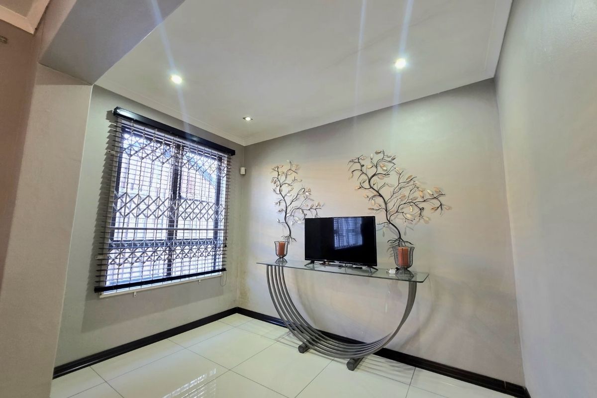A bright living area featuring neutral walls, polished floor tiles, recessed downlighting, and a window fitted with security bars and venetian blinds.