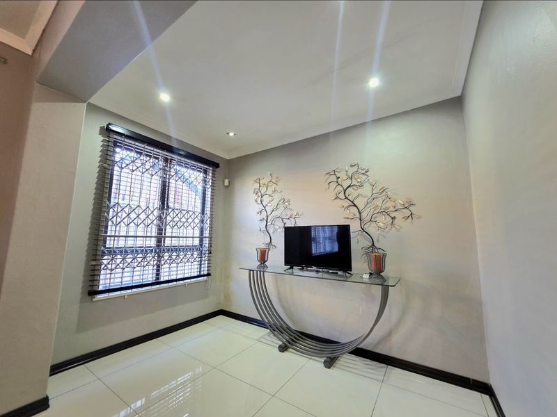 A bright living area featuring neutral walls, polished floor tiles, recessed downlighting, and a window fitted with security bars and venetian blinds.