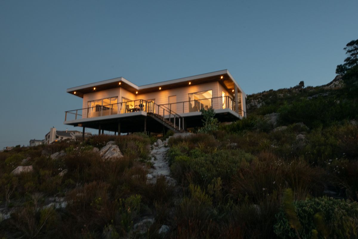 A modern elevated home featuring a spacious deck with glass balustrades, recessed lighting, and expansive sliding doors, set against a natural hillside landscape.