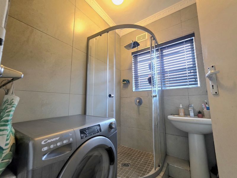 A functional bathroom featuring a corner shower enclosure, pedestal basin, and neutral wall tiling, with space for a laundry appliance.