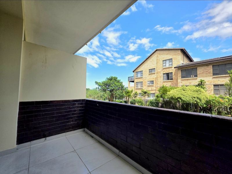 A covered balcony featuring tiled flooring and a brick balustrade, offering an open view of the surrounding complex and greenery.