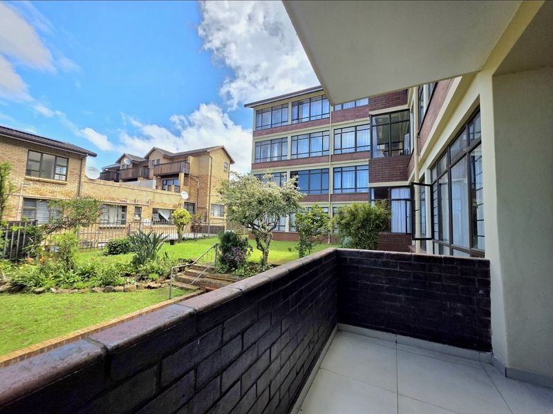 A private balcony with tiled flooring and a brick balustrade, overlooking the communal garden and surrounding apartment complex.