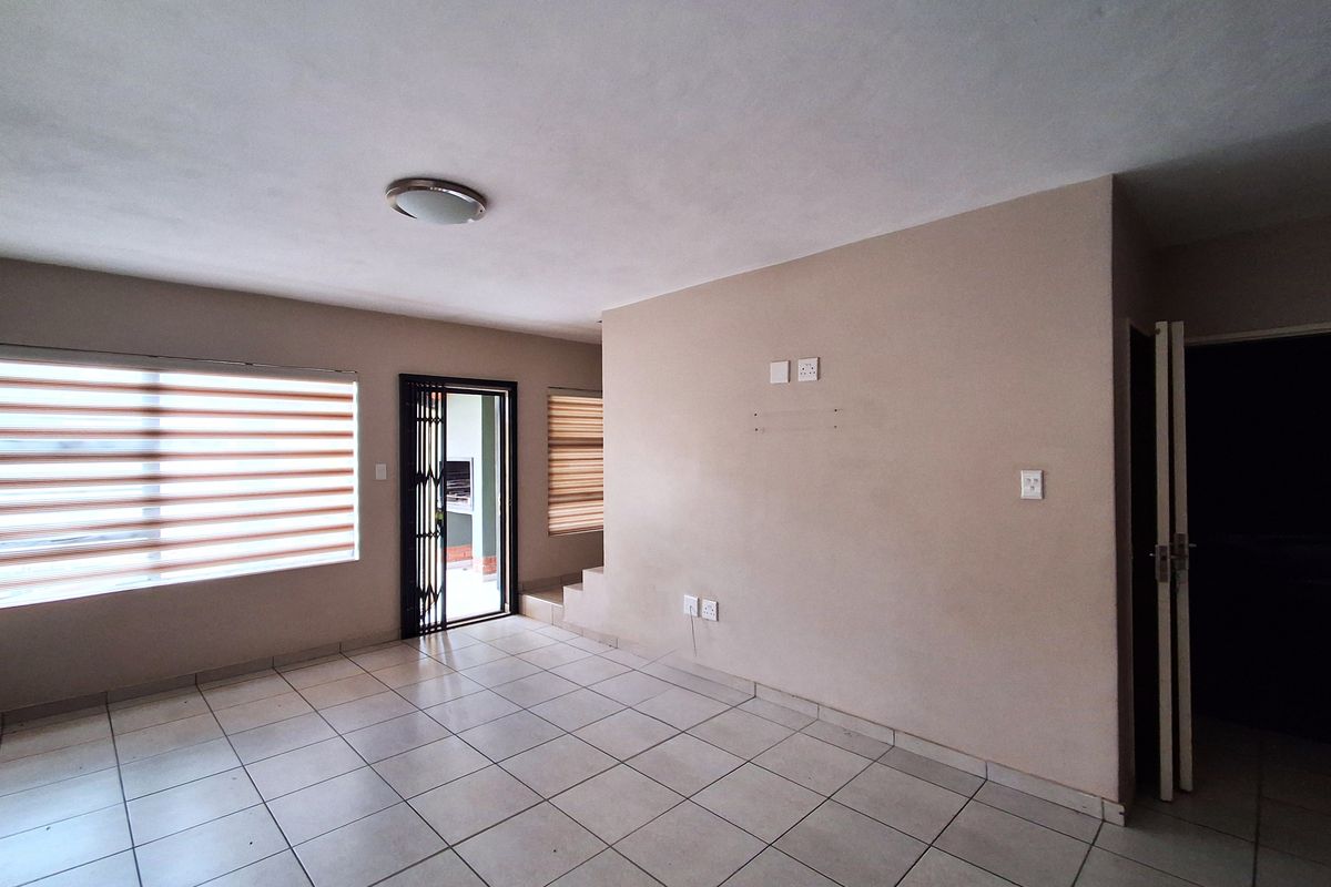 A spacious, empty living area featuring neutral tiled flooring, roller blinds, and a security gate leading to an outdoor space.