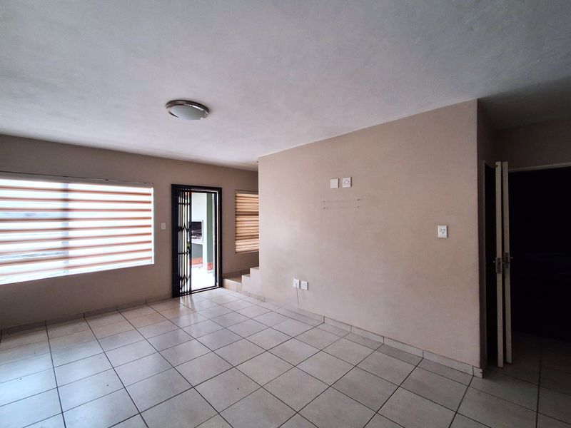 A spacious, empty living area featuring neutral tiled flooring, roller blinds, and a security gate leading to an outdoor space.