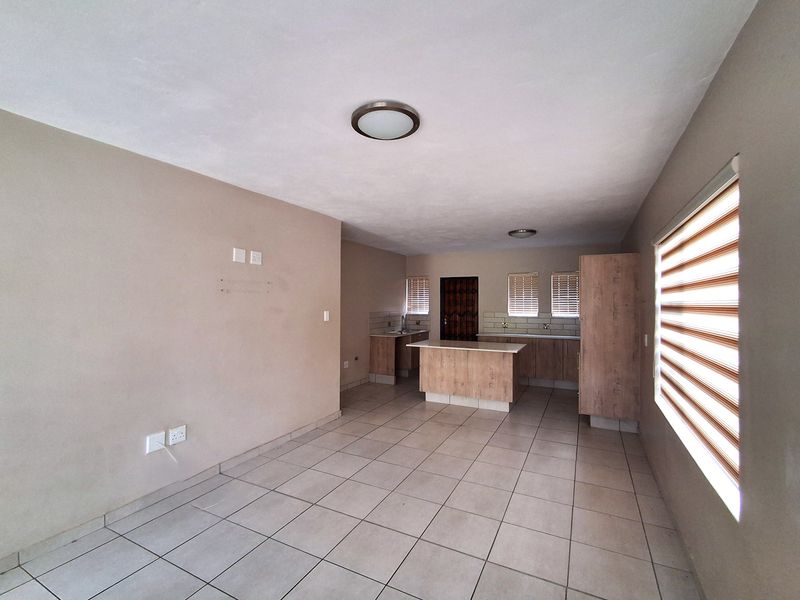Spacious open-plan living and kitchen area featuring neutral tiled flooring, modern cabinetry, a central kitchen island, and large windows with fitted blinds.