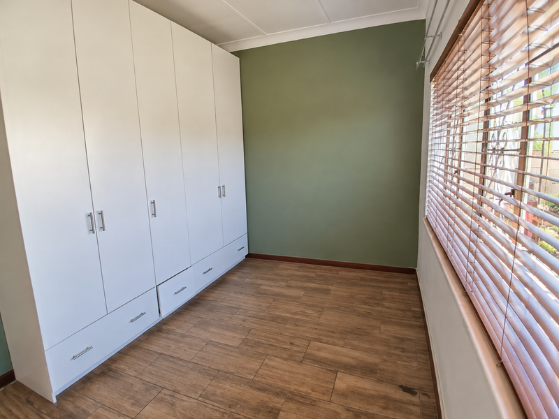 A clean, empty bedroom featuring modern white built-in cupboards, wood-look tiled flooring, and fitted wooden venetian blinds.