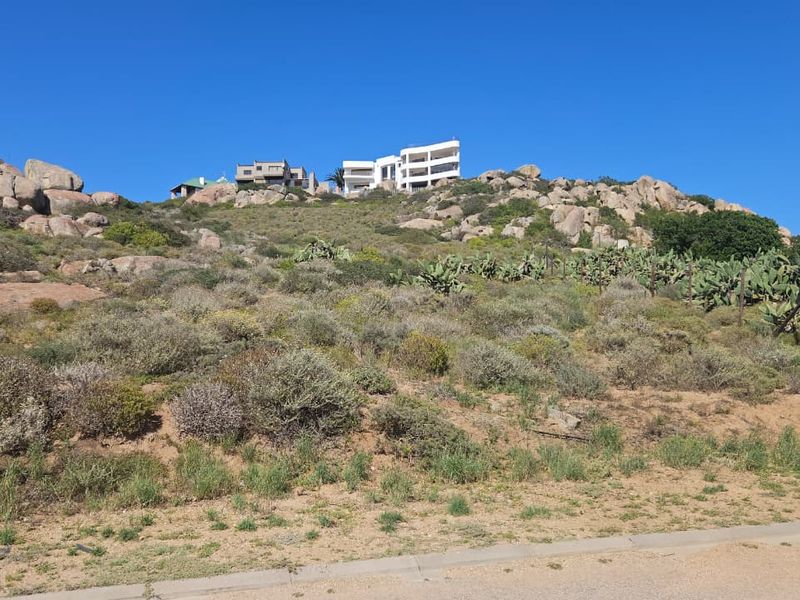 A view of a sloped, rocky residential vacant land plot with natural vegetation and surrounding hillside landscape.
