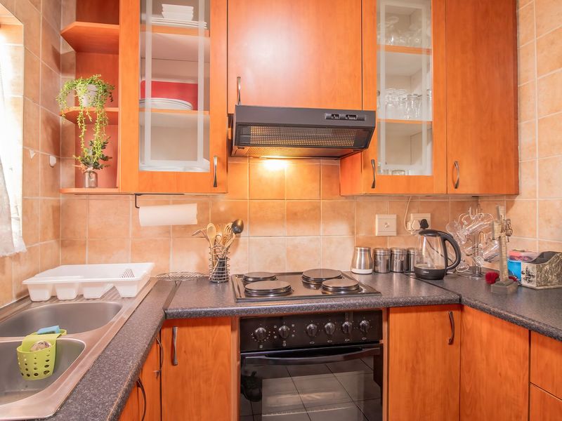 A functional kitchen featuring warm-toned wooden cabinetry, dark laminate countertops, a double stainless steel sink, and integrated electric cooking appliances with a tiled backsplash.