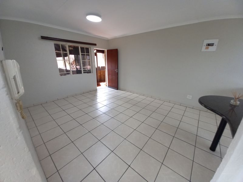 An empty, open-plan living area featuring neutral tiled flooring, a wall-mounted intercom, and a window with security bars, offering a clean, blank canvas.
