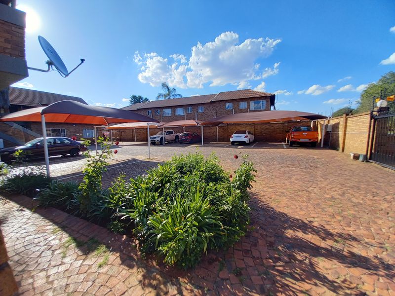 Exterior view of the apartment complex parking area featuring paved driveway, multiple shade cloth carports, and secure brick boundary walling.