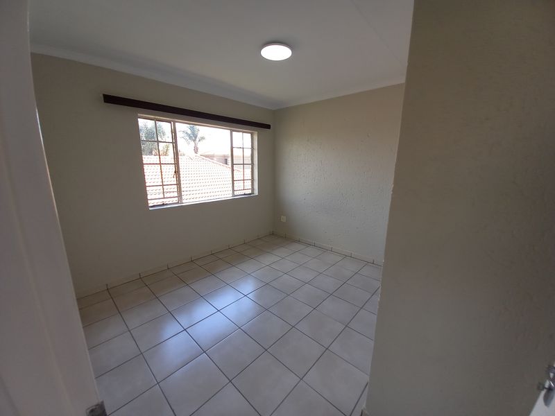 A clean, empty bedroom featuring neutral tiled flooring, a window with security bars, and a simple ceiling light fixture.