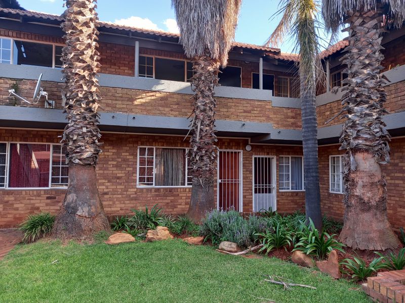 Exterior view of a face brick apartment complex featuring a shared garden area, mature palm trees, and balcony access to upper-level units.