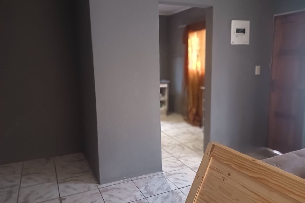 An interior view of a living area featuring tiled flooring and a wall-mounted electrical distribution board, leading into an adjacent room.