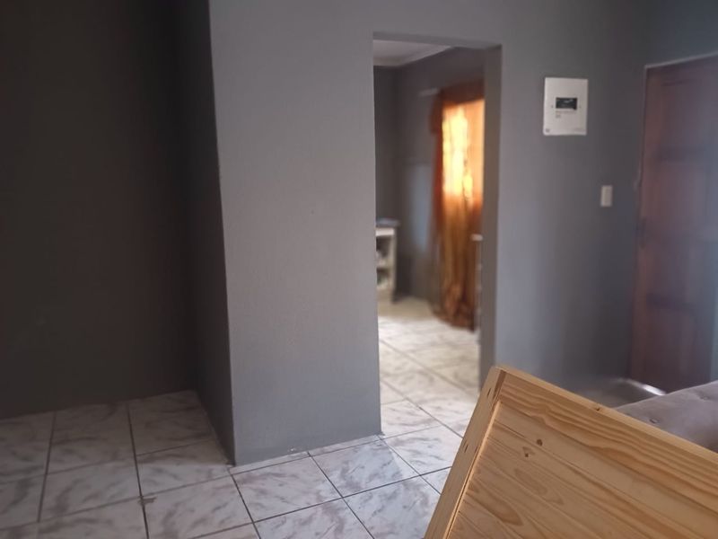 An interior view of a living area featuring tiled flooring and a wall-mounted electrical distribution board, leading into an adjacent room.