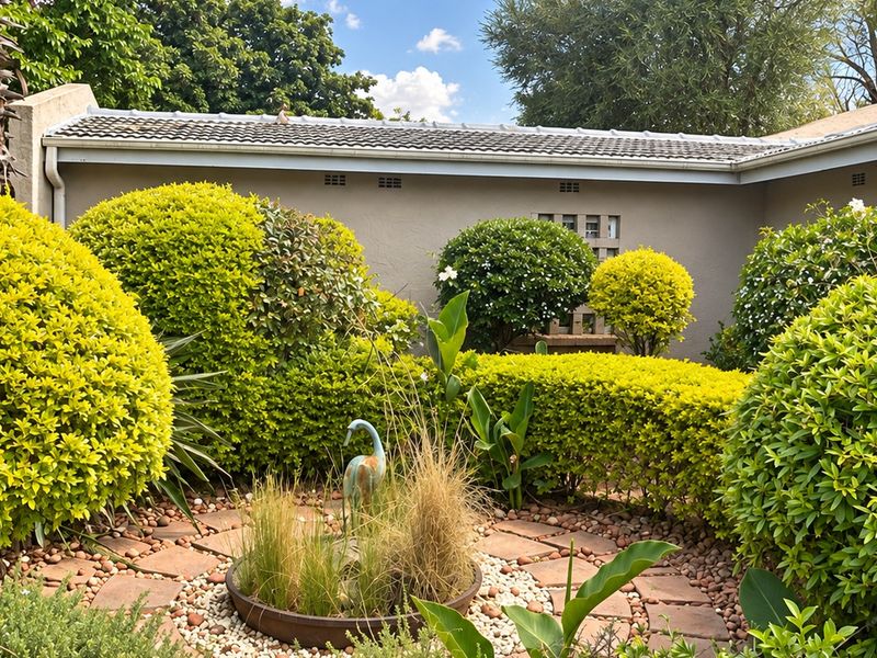 A well-maintained garden area featuring manicured shrubs, a paved stone pathway, and pebble landscaping against the exterior wall of the house.
