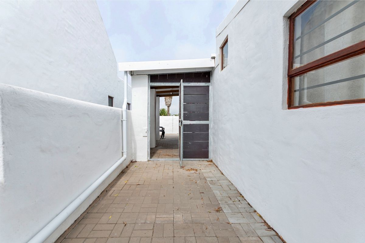 A narrow paved exterior walkway leading to a timber gate, featuring white rendered walls and a functional drainage downpipe.