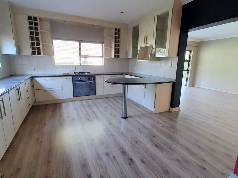 A spacious, modern kitchen featuring light-toned cabinetry, dark countertops, a built-in oven and hob, and a breakfast bar, flowing into an open-plan living area.
