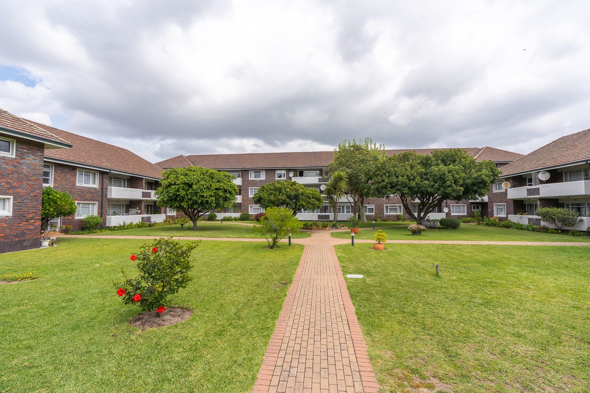A well-maintained communal garden area featuring a brick-paved walkway, mature trees, and views of the apartment building's rear facade and balconies.