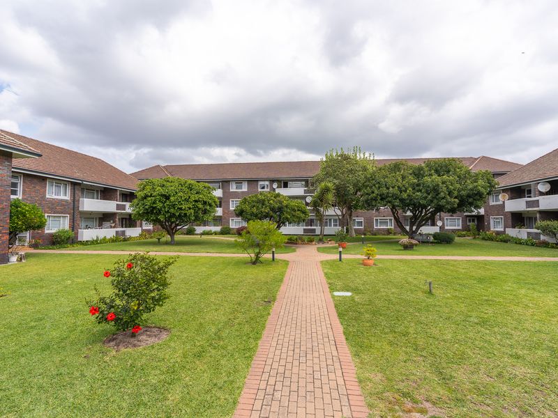 A well-maintained communal garden area featuring a brick-paved walkway, mature trees, and views of the apartment building's rear facade and balconies.