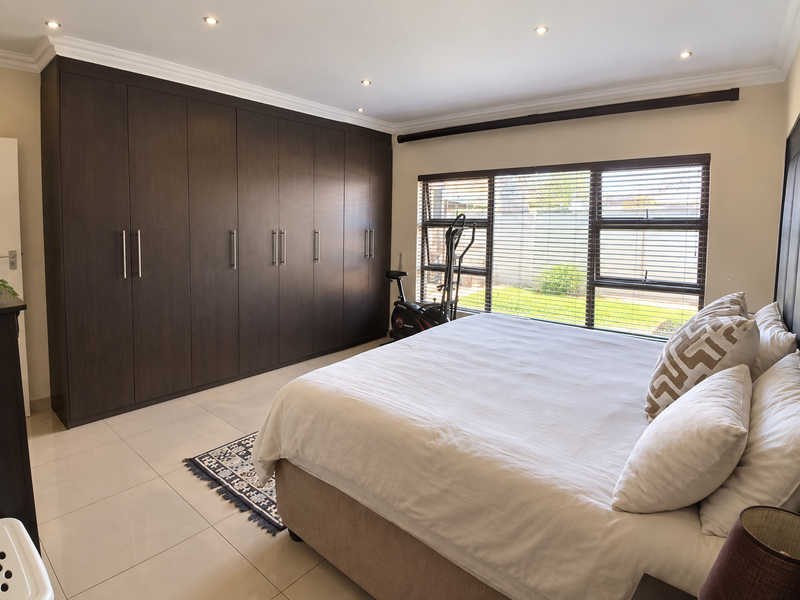 Spacious bedroom featuring extensive dark wood built-in cupboards, neutral tiled flooring, recessed lighting, and large windows with venetian blinds overlooking the garden.