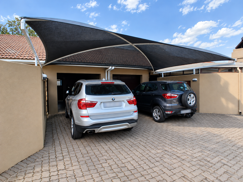 A paved driveway area featuring a modern shade sail structure providing covered parking for two vehicles in front of a double garage.