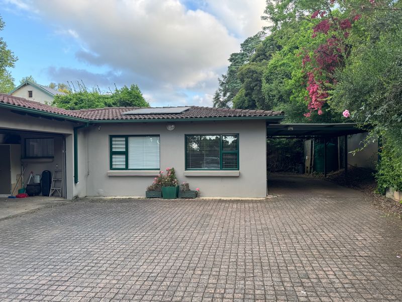 Front elevation of a cottage featuring a paved driveway, integrated garage, carport, and established garden surroundings.
