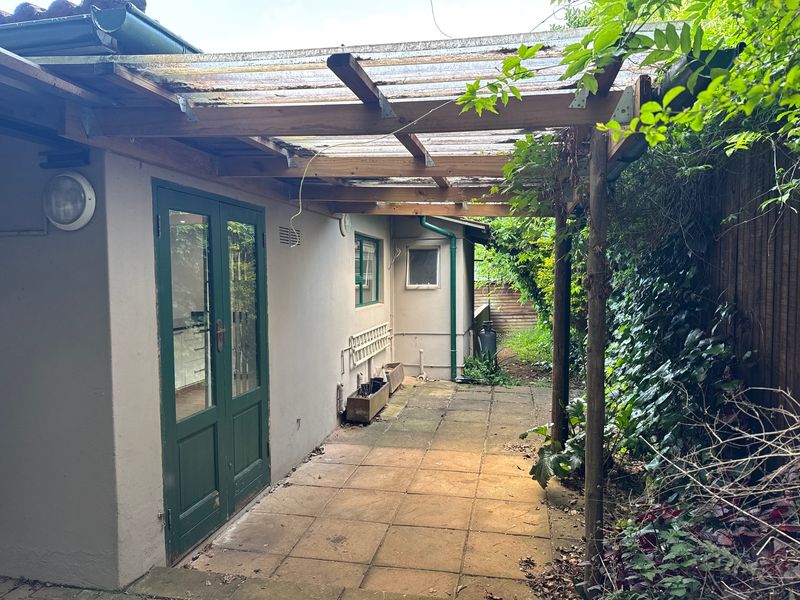 A covered outdoor patio area featuring paved flooring, a timber pergola with translucent roofing, and direct access to the interior via green french doors.