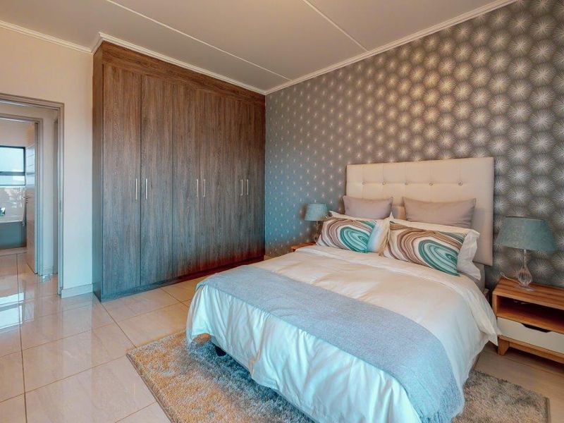 A well-maintained bedroom featuring built-in wooden cupboards, tiled flooring, and a decorative wallpaper feature wall, with an adjoining bathroom visible.