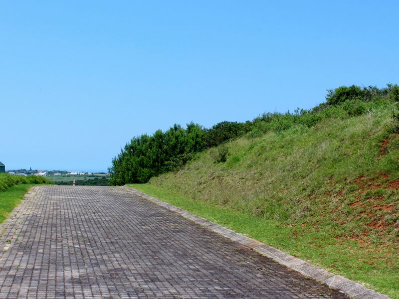 A paved driveway leading through a residential area with a sloped grass embankment and distant coastal views.