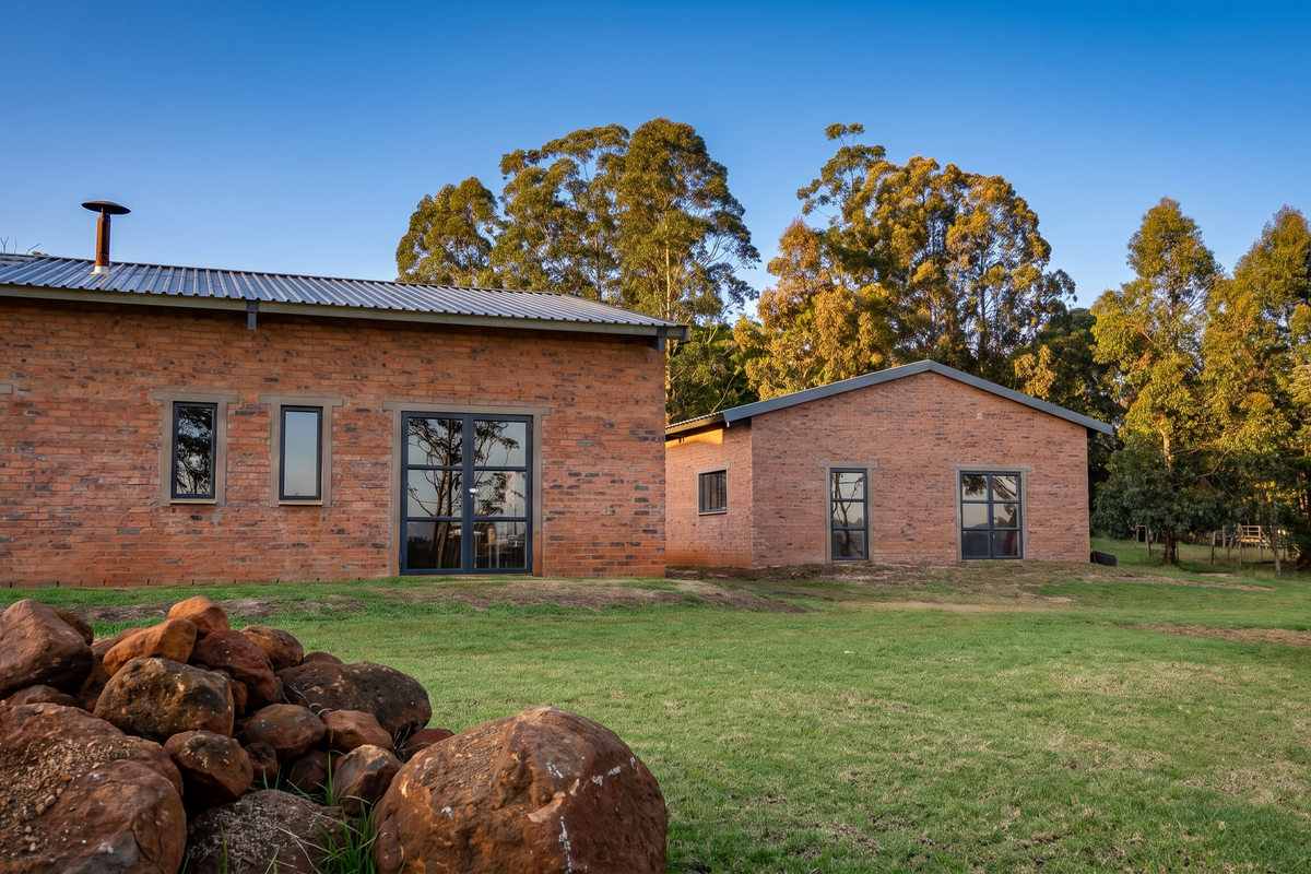 Rear view of a modern industrial-style brick dwelling featuring large glass doors, aluminium windows, and a corrugated metal roof set in a landscaped garden.