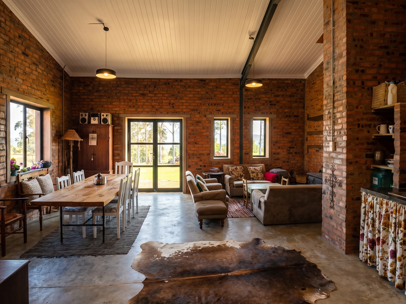 Spacious open-plan living and dining area featuring rustic exposed brick walls, vaulted ceilings, concrete floors, and direct access to the garden through double doors.