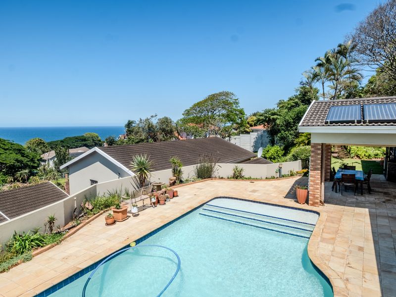 Outdoor pool area featuring a tiled patio, covered entertainment space, and solar water heating panels, set against a scenic coastal view.