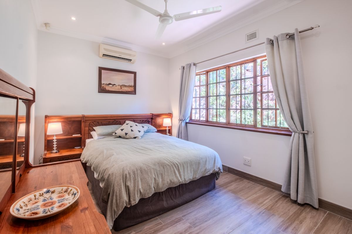 A well-maintained bedroom featuring wood-look tile flooring, a ceiling fan, split-system air conditioning, and timber-framed windows overlooking a garden area.