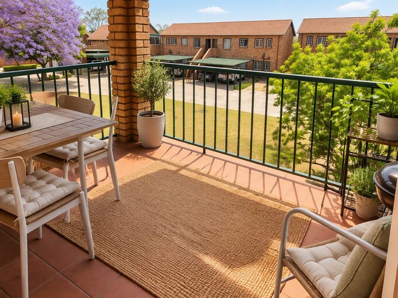 A well-maintained covered balcony featuring tiled flooring, a brick support pillar, and metal railings, overlooking the complex grounds.