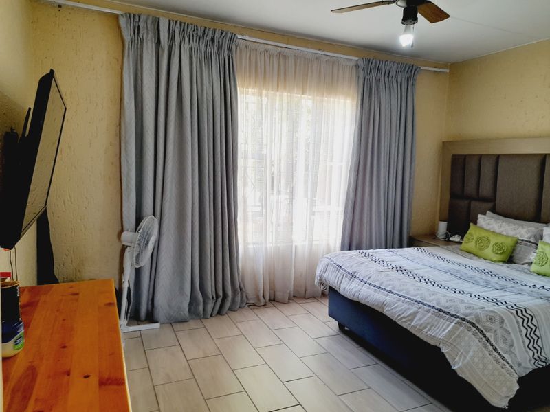 A well-lit bedroom featuring tiled flooring, a ceiling fan, and large windows dressed with floor-to-ceiling curtains.