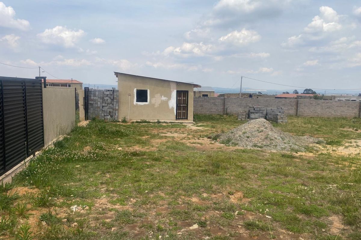 A view of a residential plot featuring a small outbuilding, perimeter walling, and a metal gate, with unfinished ground and construction materials present.