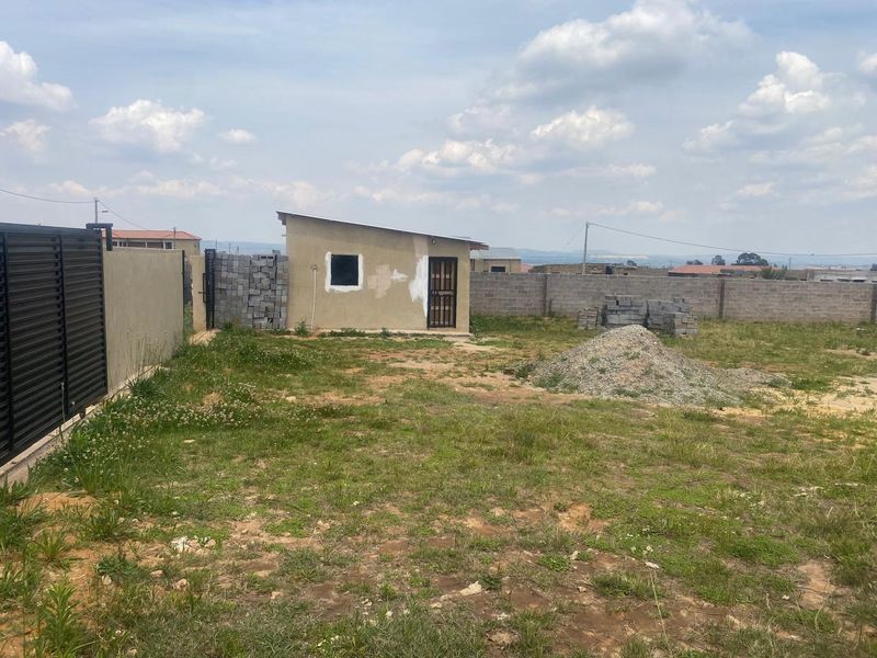 A view of a residential plot featuring a small outbuilding, perimeter walling, and a metal gate, with unfinished ground and construction materials present.