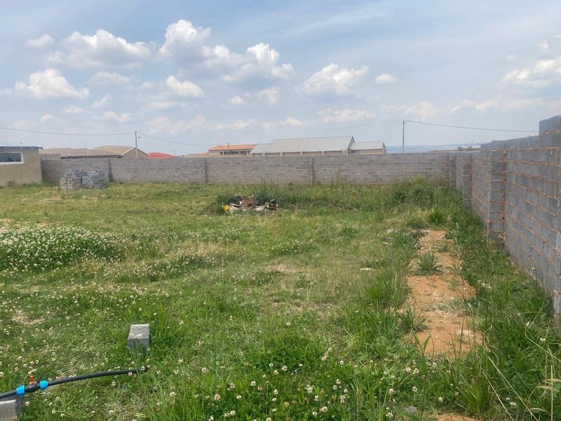 A vacant residential plot enclosed by concrete block perimeter walls, featuring open grassed space and surrounding neighborhood views.